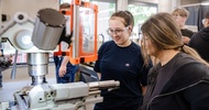 Zwei Personen stehen vor einer Werkzeugmaschine mit transparentem Schutzschild in einer Werkstatt. Beide tragen Schutzbrillen und blicken auf den Arbeitsbereich der Maschine. Im Hintergrund befinden sich weitere Personen