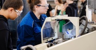 Seitliche Aufnahme von zwei Personen, die nebeneinander an einer Maschine mit Schutzabdeckung stehen. In der Maschine ist ein Metallstück eingespannt