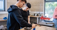 Zwei Personen stehen an einer Werkbank in einer Werkstatt. Auf der Arbeitsfläche liegen ein Hammer, Metallstifte und weitere Werkzeuge