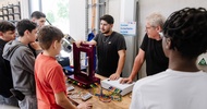 Mehrere Personen stehen um eine Werkbank mit elektronischen Komponenten und Kabelverbindungen. Zwei Personen stehen an einer technischen Anwendung mit einem Türrahmen-Modell und angeschlossenen Geräten. Im Hintergrund hängt ein Schild mit der Aufschrift „Station Sicherheitstechnik“