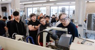 Mehrere Personen stehen in einer Werkstatt um eine technische Maschine mit Schutzabdeckung. Eine Person mit Schutzbrille bedient die Maschine, während die Gruppe den Arbeitsvorgang beobachtet
