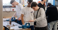 Mehrere Personen arbeiten in einer Werkstatt an Werkbänken mit Schraubstöcken und Werkzeugen. Im Vordergrund bearbeitet eine Person ein eingespanntes Metallstück mit einer Feile. Weitere Personen stehen an benachbarten Arbeitsplätzen. Auf der Werkbank liegen Werkzeuge und Materialien