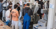 Mehrere Personen stehen in einer Werkstatt um eine Fräsmaschine und beobachten bzw. bedienen die Anlage. Eine Person in blauer Arbeitskleidung arbeitet direkt an der Maschine. Im Vordergrund befinden sich Werkzeuge und ein Schraubstock auf einer Werkbank