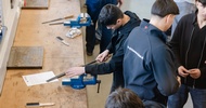 Mehrere Personen stehen an einer langen Werkbank in einer Werkstatt. Eine Person bearbeitet ein eingespanntes Metallteil mit einer Feile in einem blauen Schraubstock. Auf der Werkbank liegen weitere Feilen, Werkzeuge und Metallteile