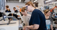 Mehrere Personen arbeiten in einer Werkstatt an einer Maschine. Zwei Personen im Vordergrund tragen Schutzbrillen und bedienen gemeinsam eine Anlage