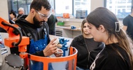 Eine Person mit Schutzbrille und Arbeitskleidung hält ein kleines Werkstück neben einer Maschine mit transparentem Schutzschild. Ihr gegenüber stehen zwei weitere Personen. Eine der Personen blickt auf das Werkstück. Im Hintergrund befinden sich Werkbänke, Schraubstöcke und weitere Personen