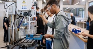 Mehrere Personen stehen an einer Fräsmaschine in einer Werkstatt. Zwei Personen bedienen die Maschine und halten Werkstück sowie Bedienelemente. Beide tragen Schutzbrillen. Auf dem Arbeitstisch der Maschine liegen Metallspäne