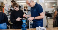 Zwei Personen stehen an einer Werkbank in einer Werkstatt. Am Tisch ist ein Schraubstock, in dem ein Werkstück steckt. Beide Personen blicken auf den Schraubstock. Im Hintergrund befinden sich Maschinen und weitere Arbeitsplätze