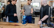 Drei Personen stehen an einer Werkbank in einer Werkstatt. Eine Person spricht und gestikuliert, während eine weitere Person ein Werkzeug hält. Auf der Werkbank befinden sich ein Schraubstock, Werkzeuge und Messgeräte