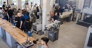 Aufnahme eines großen Raums aus der Vogelperspektive mit mehreren Maschinen, Werkbänken und Arbeitsstationen. Zahlreiche Personen arbeiten oder sprechen an unterschiedlichen Arbeitsplätzen. Im Vordergrund steht eine lange Werkbank mit Schraubstöcken, Werkzeugen und Messgeräten