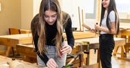 An einem Werktisch steht eine jugendliche Person, die ein dünnes, eingespanntes Holzbrett mit einem Schraubenzieher bearbeitet. Im Hintergrund steht eine weitere jugendliche Person