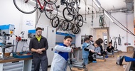 In einem großen Raum stehen vier jugendliche an einem Tisch mit Drehbänken und probieren diese aus. Eine Person mit Schutzbrille steht im Hintergrund. Von der Decke hängen Fahrräder<br />
<br />
