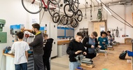 In einem großen Raum stehen vier jugendliche an einem Tisch mit Drehbänken und probieren diese aus. Zwei weitere Kinder und eine Person mit Schutzbrille stehen an einem Gerät. Von der Decke hängen Fahrräder