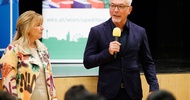 Frontalaufnahme zweier nebeneinander stehender Personen. Die rechte Person spricht in ein Mikrofon. Ihnen gegenüber sitzen mehrere Personen auf Stühlen