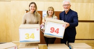 Porträt von drei nebeneinander stehender Personen. Zwei Personen halten einen Karton, der in braunes Papier eingepackt ist. Auf dem linken steht 34, auf dem rechten 94