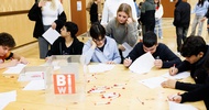 Um einen großen Tisch sitzt eine Gruppe von Teenagern. Jeder Teenager hat einen Zettel vor sich und schreibt darauf. Zwei Personen stehen neben dem Tisch