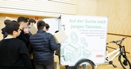 Hinter einem geschlossenen Anhänger, der an einem Fahrrad montiert ist, steht eine Gruppe von Personen. Ein paar von ihnen halten übereinander gestapelte Kartons