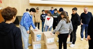 Am Boden stehen mehrere, in braunem Papier verpackte, rechteckige Objekte. Um diese steht eine Gruppe von Teenagern. Ein paar blicken auf die Objete