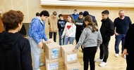 Am Boden stehen mehrere, in braunem Papier verpackte, rechteckige Objekte. Um diese steht eine Gruppe von Teenagern. Ein paar blicken auf die Objete