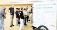 Hinter einem geschlossenen Anhänger, der an einem Fahrrad montiert ist, steht eine Gruppe von Personen. Ein paar von ihnen halten übereinander gestapelte Kartons