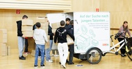 Hinter einem geschlossenen Anhänger, der an einem Fahrrad montiert ist, stehen sieben Personen, die alle einen Stapel Kartons halten