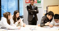 An einem Tisch sitzen vier Jugendliche in zwei Gruppen jeweils zu zweit. Beide Gruppen halten Stifte und schreiben Zettel. Am Tisch steht eine Person in einem Anzug, die den Jugendlichen zusieht