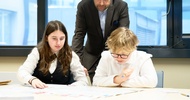Zwei Kinder sitzen nebeneinander an einem großen Tisch. Auf dem Tisch liegen verschiedene Zettel, auf die sie blicken. Hinter ihnen steht eine weitere Person, die auf die Zettel blickt