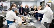 In einer Gastronomieküche stehen um einen Arbeitsblock zwei Personen mit Haarnetzen und Schürzen, die Jugendlichen, die neben ihnen stehen etwas erklären. Im Hintergrund sind weitere Jugendliche sowie Erwachsene, die sich unterhalten