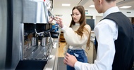 Neben einer Kaffeemaschine stehen eine jugendliche Person sowie ein Kellner. Die jugendliche Person drückt einen Knopf auf der Maschine