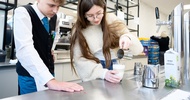 Zwei Jugendliche stehen in einer Gastroküche. Die rechte Person leert aus einer Milchkanne Kaffee in ein Glas mit Milchschaum. Die Person daneben schaut zu und trägt eine dunkle Weste sowie eine Krawatte. Auf dem Tresen ist ein Zapfhahn