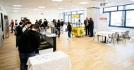 In einem großen Raum sind mehrere Tische, ein Tresen, hinter dem Personen stehen, eine Plakatwand, sowie viele Personen, die in Gruppen oder alleine stehen. Zwischen den Personen sind Kellner:innen