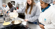 Eine Person mit Haarnetz und Schürze sowie eine Jugendliche stehen in einer Gastronomieküche neben einer Herdplatte. Die jugendliche Person hält eine Pfanne, in die sie aus einer Schüssel eine helle Flüssigkeit kippt