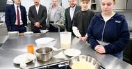 Neben einem Arbeitstisch in einer Gastronomieküche stehen vier erwachsene Personen sowie zwei Jugendliche. Auf dem Arbeitstisch sind Schüsseln, Teller, ein Topf und eine Pfanne mit einem hellen Teig