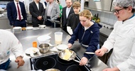 Neben einem Arbeitstisch in einer Gastronomieküche stehen vier erwachsene Personen. Neben ihnen sind zwei Jugendliche sowie eine Person mit Haube und Schürze. Auf dem Arbeitstisch ist eine Herdplatte, auf der eine Pfanne steht. Eine der jugendlichen Personen kocht darin