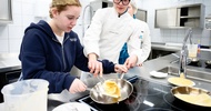 In einer Gastronomieküche stehen neben einer Herdplatte. Eine jugendliche Person sowie eine Person mit Haube, Brille und Schürze. Auf der Herdplatte steht eine Pfanne. Die jugendliche Person und die andere Person kochen darin gemeinsam ein Omlette