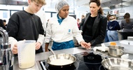 In einer Gastronomieküche stehen neben einer Herdplatte eine jugendliche Person, eine Person mit Haarnetz und Schürze sowie eine erwachsene Person. Auf der Herdplatte steht eine Pfanne. Die jugendliche Person hält ein Plastikgefäß, in dem eine helle Flüssigkeit ist