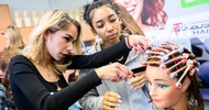 Ein Modepuppenkopf hat die Haare eingedreht. Neben dem Kopf steht eine erwachsene Person, die eine Haarsträhne und einen Kamm hält. Daneben steht eine jugendliche Person, die zusieht
