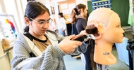 Seitliche Aufnahme des Hinterkopfes einer Modepuppe, die oben keine Haare hat. Im unteren Bereich sind Haare. Eine Person hält einen Rasierer und einen Kamm auf die Haare