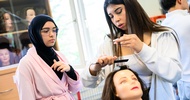 Eine Person hält die Haarsträhne eines Modepuppenkopfes mit der linken Hand. Mit der rechten Hand kämmt sie die Strähne. Neben ihr steht eine jugendliche Person, die zusieht