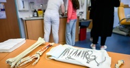 Im Vordergrund liegen auf einem Tisch verschiedene Utensilien wie Scheren oder Pinzetten. Daneben liegen Modelle von Händen und Knochen. Im Hintergrund stehen Personen herum