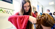 Ein Modepuppenkopf mit langem Haar ist auf einer Metallstange montiert. Dahinter steht eine jugendliche Person, die die Haare der Puppe mit einem Lockenstab in Form bringt
