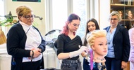 Ein Modepuppenkopf mit langem Haar ist auf einer Metallstange montiert. Dahinter steht eine jugendliche Person, die die Haarsträhnen mit Handschuhen bunt färbt. Daneben stehen weitere Personen