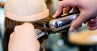Nahaufnahme des Hinterkopfes einer Modepuppe, die oben keine Haare hat. Im unteren Bereich sind Haare. Eine Person hält einen Rasierer und einen Kamm auf die Haare