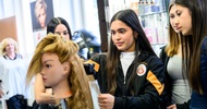 Ein Modepuppenkopf mit langem Haar ist auf einer Metallstange montiert. Dahinter steht eine jugendliche Person, die die Haare der Puppe glättet. Daneben stehen weitere jugendliche Personen