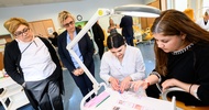 In einem Raum sind neben einem kleinen, weißen Tisch mit einer Lampe zwei Personen. Die linke Person trägt einen Kittel. Auf dem Tisch liegen verschiedene Zettel