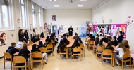 In einem großen Raum sitzen viele jugendliche Personen an Tischen. Alle blicken zu einer Person, die ihnen gegenüber steht und gerade spricht