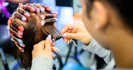 Ein Modepuppenkopf hat die Haare eingedreht. Neben dem Kopf steht eine Person, die eine Haarsträhne auf einen Lockenwickler aufdreht