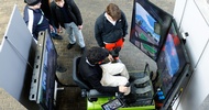 Eine jugendliche Person sitzt auf einem Simulator eines Gabelstaplers. Gegenüber der Person sind drei Bildschirme. Neben der Person stehen vier weitere Personen