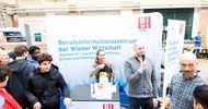 An einem hohen Tisch stehen zwei Personen. Die rechte Person hält ein Mikrofon vor den Mund. Hinter ihnen ist ein großer Banner, auf dem Berufsinformationszentrum der Wiener Wirtschaft steht. Um den Tisch stehen weitere jugendliche und erwachsene Personen