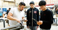 Eine erwachsene Person und zwei Jugendliche stehen neben neben einem Tisch auf einer Messe. Auf dem Tisch sind verschiedene Werkzeuge zur Metallbearbeitung. Die Person hält ein Werkzeug